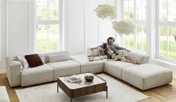 Großzügiges, helles Wohnzimmer mit bodentiefen Fenstern und modernem, beigem Ecksofa. Ein Paar sitzt entspannt auf dem Sofa und genießt gemeinsam ein Buch.